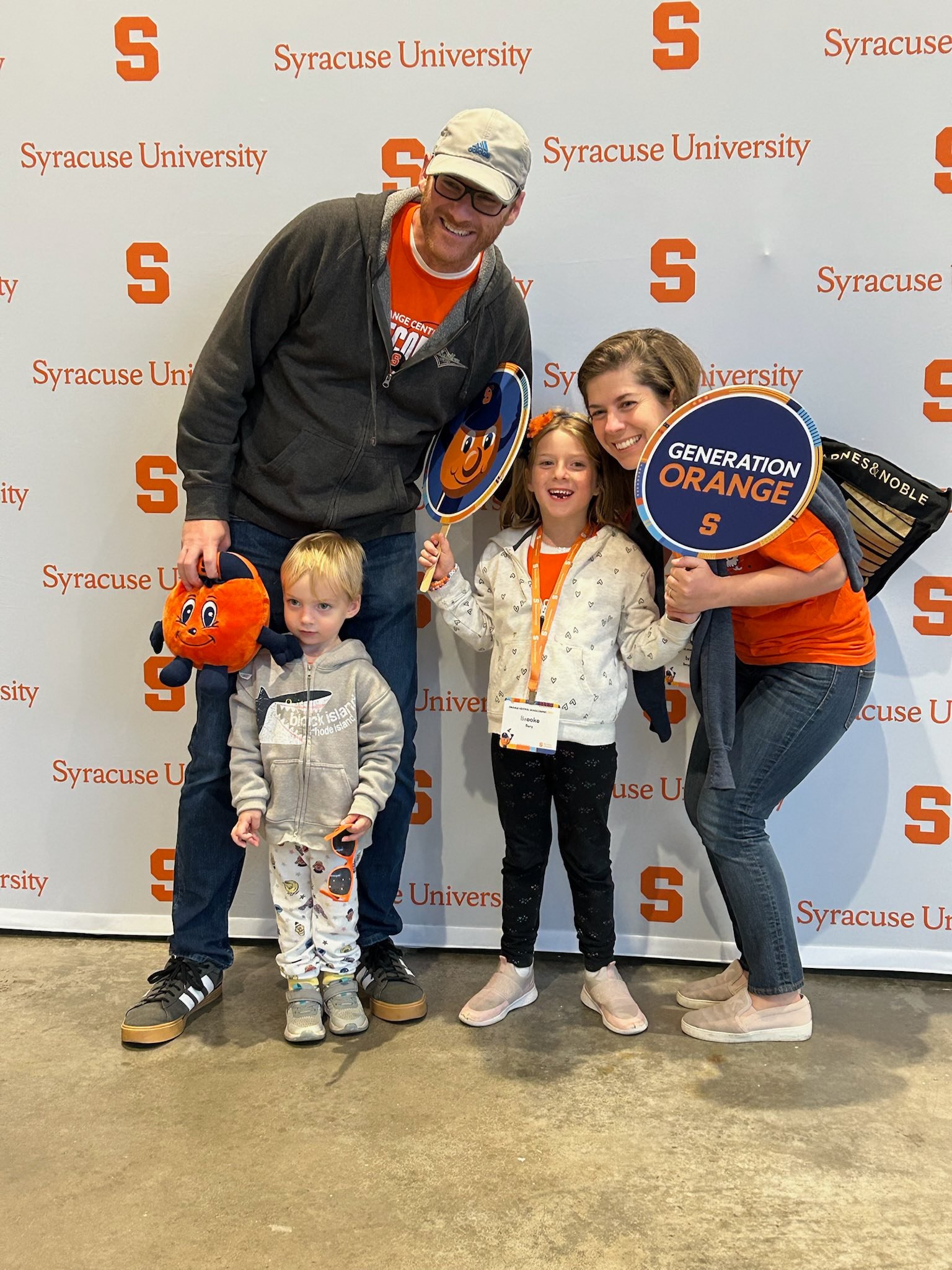 Family of four posing at photo backdrop holding Syracuse-themed props including Otto mascot sign and "Generation Orange" paddle