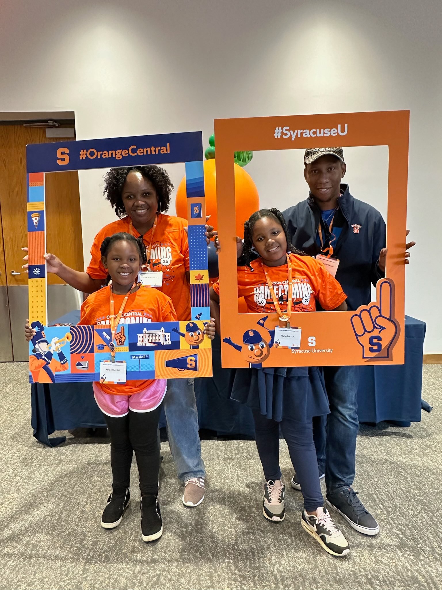 Family with two young daughters in Syracuse Homecoming t-shirts holding decorative photo frames at event