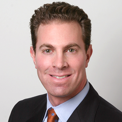 Professional headshot of a man in a dark suit and patterned tie, smiling at the camera against a light background