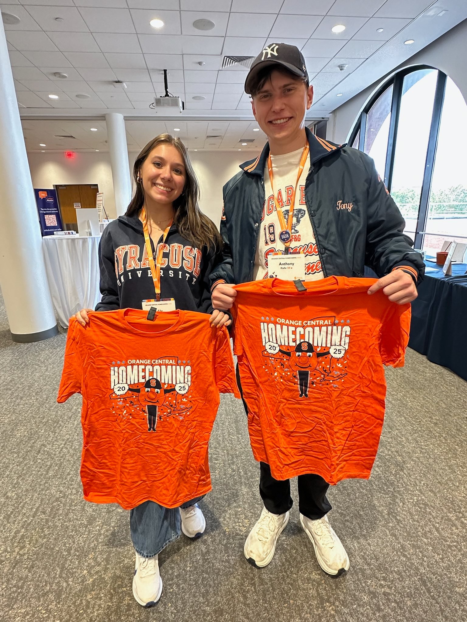 Two people displaying orange Homecoming 2025 t-shirts featuring illustrated Otto mascot design in campus building