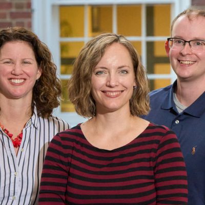 Three smiling professionals pose together in front of a brick building with illuminated windows.