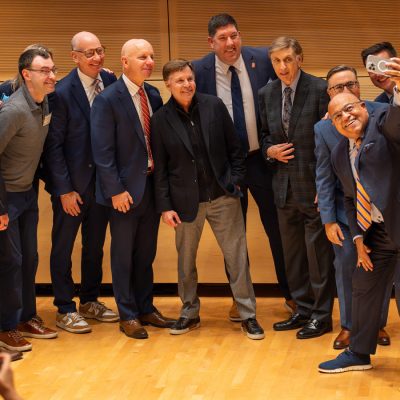 Group of people dressed in business attire standing close together on a wooden stage, posing for a selfie. One person in the front holds a phone extended outward to capture the photo