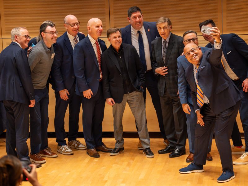 Group of people dressed in business attire standing close together on a wooden stage, posing for a selfie. One person in the front holds a phone extended outward to capture the photo
