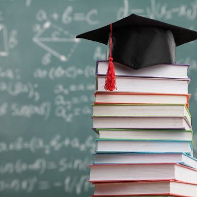 A black graduation cap with a red tassel rests on top of a tall stack of colorful textbooks, with a chalkboard covered in mathematical equations visible in the blurred background