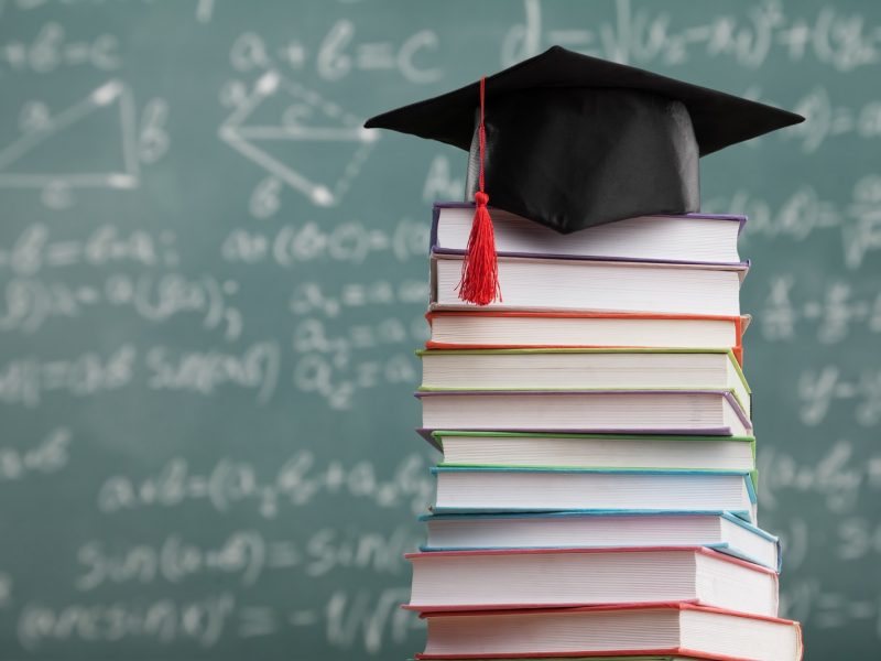 A black graduation cap with a red tassel rests on top of a tall stack of colorful textbooks, with a chalkboard covered in mathematical equations visible in the blurred background