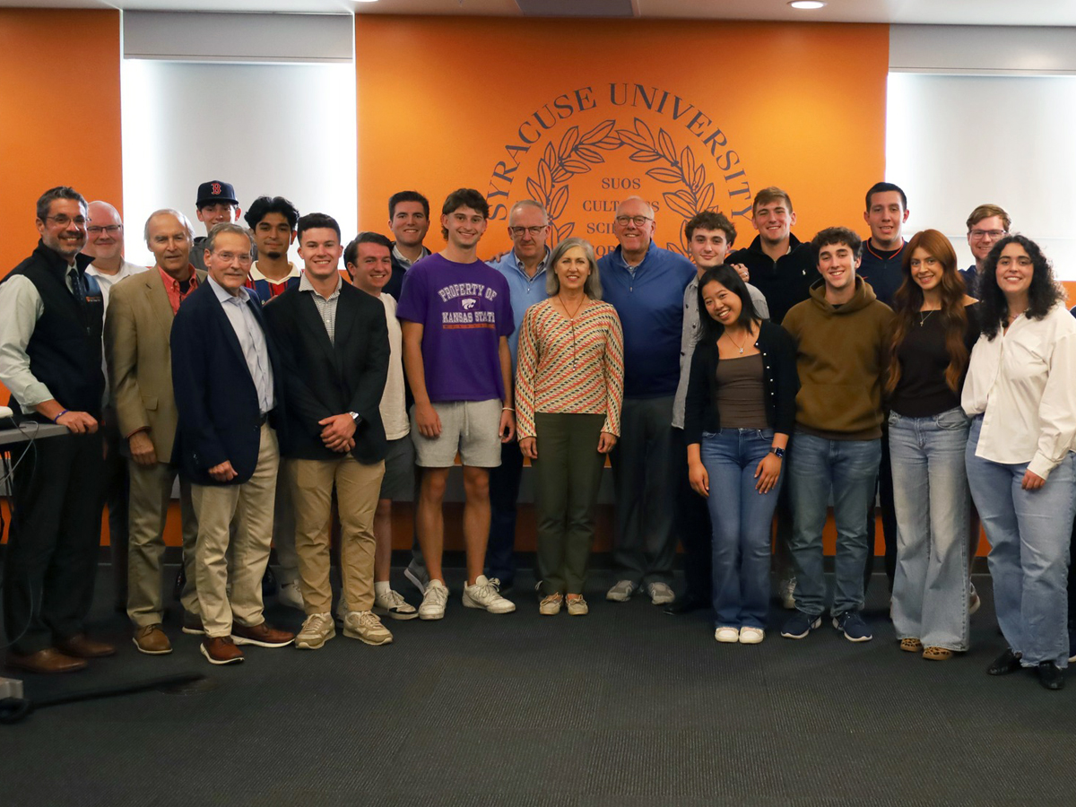 A large group of approximately 20-25 people, including students and faculty members, standing together for a group photo in front of an orange wall featuring the Syracuse University seal and motto at the top.