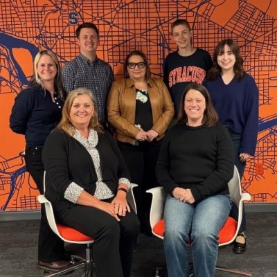 Seven people pose in front of an orange wall featuring a blue map. Two people are seated in the front row and five people are standing behind them. From left to right in the back: a person in a navy top, a person in a blue plaid shirt, a person in a tan cardigan, a person in a Syracuse sweatshirt and a person in a navy blue top. The two seated individuals wear black tops, one with a black and white patterned collar, the other in a black sweater with jeans.