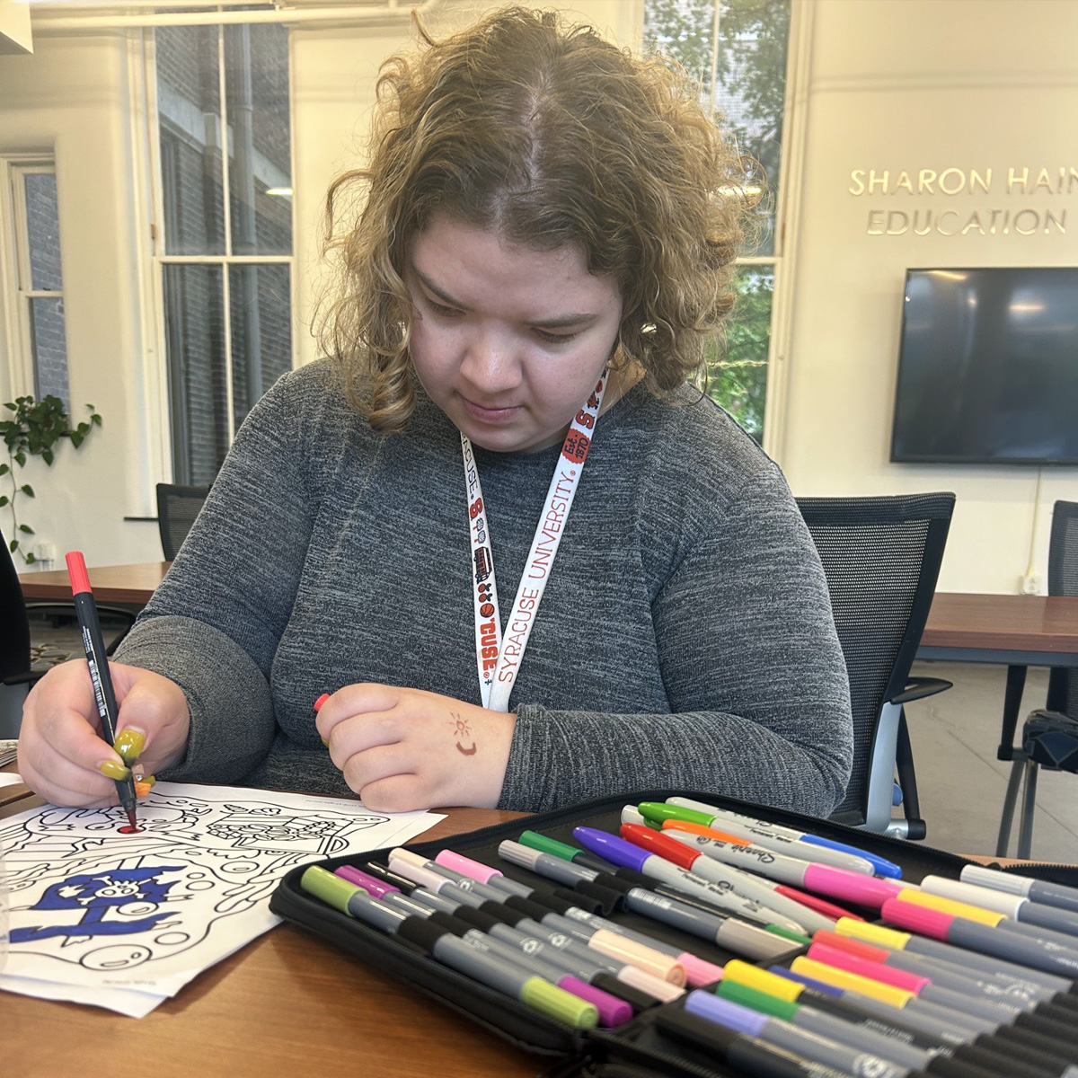 A student wearing a Syracuse University lanyard concentrates on coloring an intricate mandala design with markers at a desk in an educational setting.