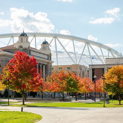 Historic campus building with fall foliage and stadium in background under partly cloudy sky