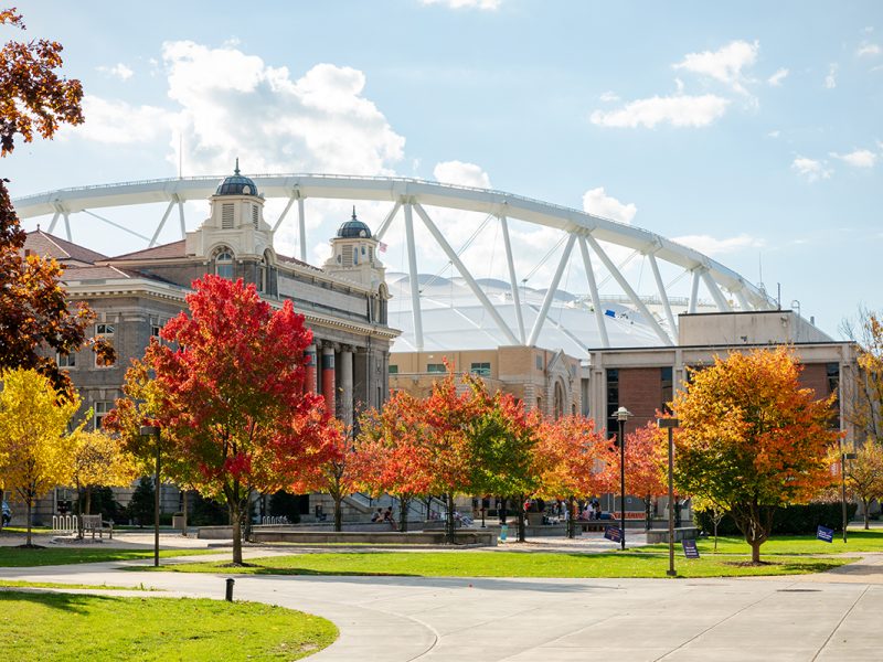 Historic campus building with fall foliage and stadium in background under partly cloudy sky