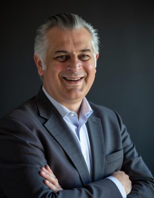 Person wearing a dark suit jacket over a light blue dress shirt, standing with arms crossed against a dark background.