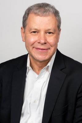 Person wearing a black suit jacket over a white dress shirt against a plain light background.
