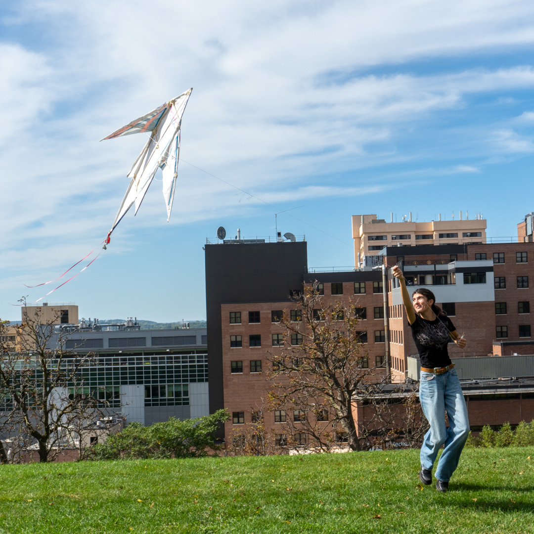 Person flying a large, colorful kite in a grassy field with urban buildings in the background under a partly cloudy sky