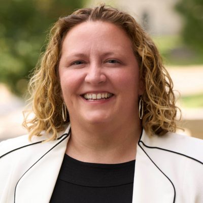 Person with curly hair in a white blazer smiling outdoors in front greenery.