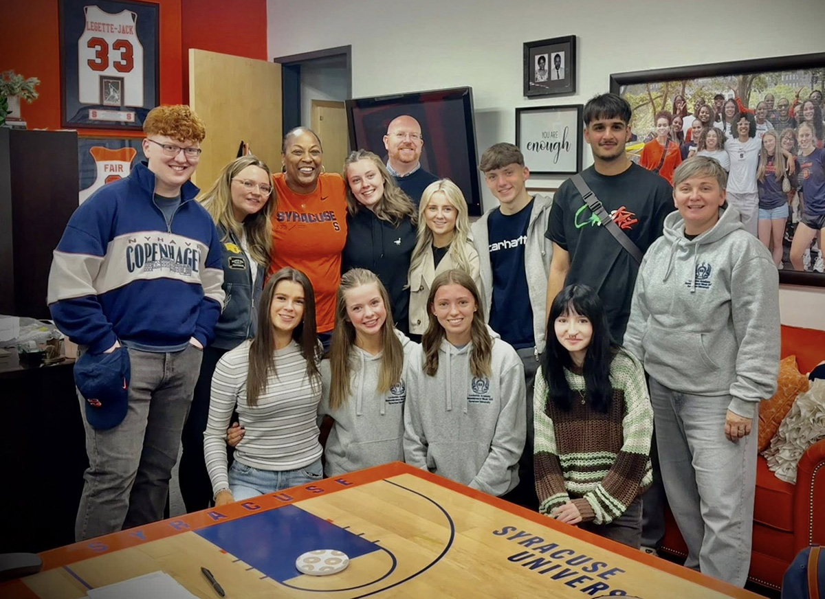 Group of thirteen people posing around a Syracuse University table in a decorated room with framed photos and a sports jersey