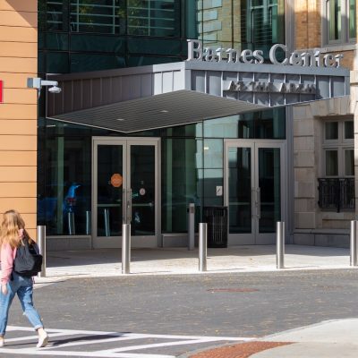Person crossing a street toward the entrance of the Barnes Center at The Arch, a modern building with glass doors and tan panels.