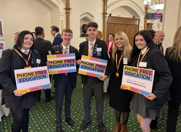 Five young people in school uniforms and formal wear hold "Phone Free Education" signs while standing together in an ornate hall with white arched walls and green patterned carpet. They wear orange visitor lanyards and smile at the camera.