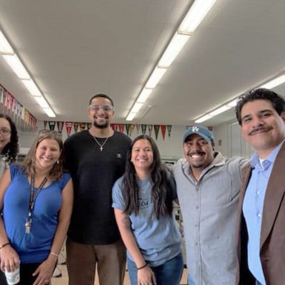 Six people smiling in a classroom decorated with college pennants.