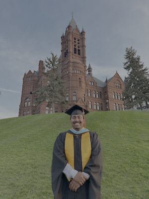 Person in graduation attire standing on a grassy hill with a historic red-brick building and turrets in the background.