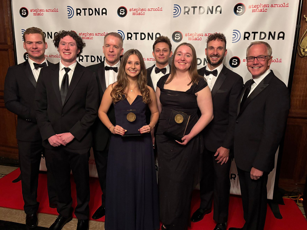 Eight people in formal attire pose on a red carpet, with two women in the center holding award plaques, flanked by six men in tuxedos. Backdrop displays RTDNA and Stephen Arnold Music logos.