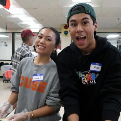 Two student volunteers wearing name tags smile while preparing food at a community service event in a cafeteria-style room with other volunteers in the background.