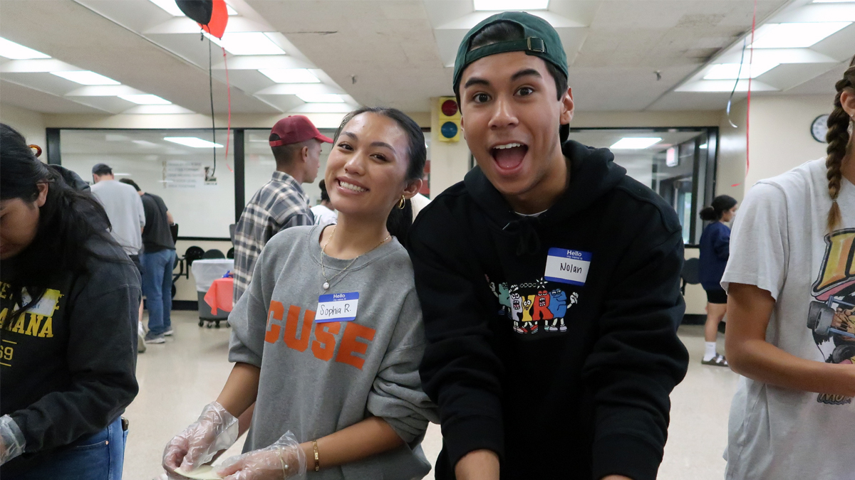 Two student volunteers wearing name tags smile while preparing food at a community service event in a cafeteria-style room with other volunteers in the background.