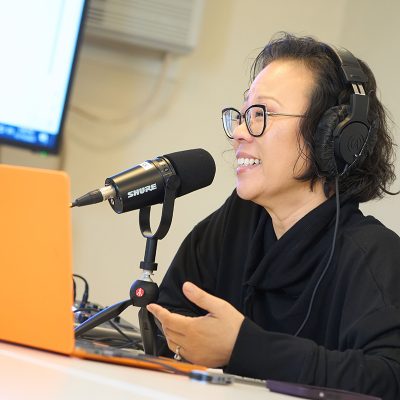 Person speaking into a professional microphone while wearing headphones, seated at a desk with an orange laptop and audio equipment