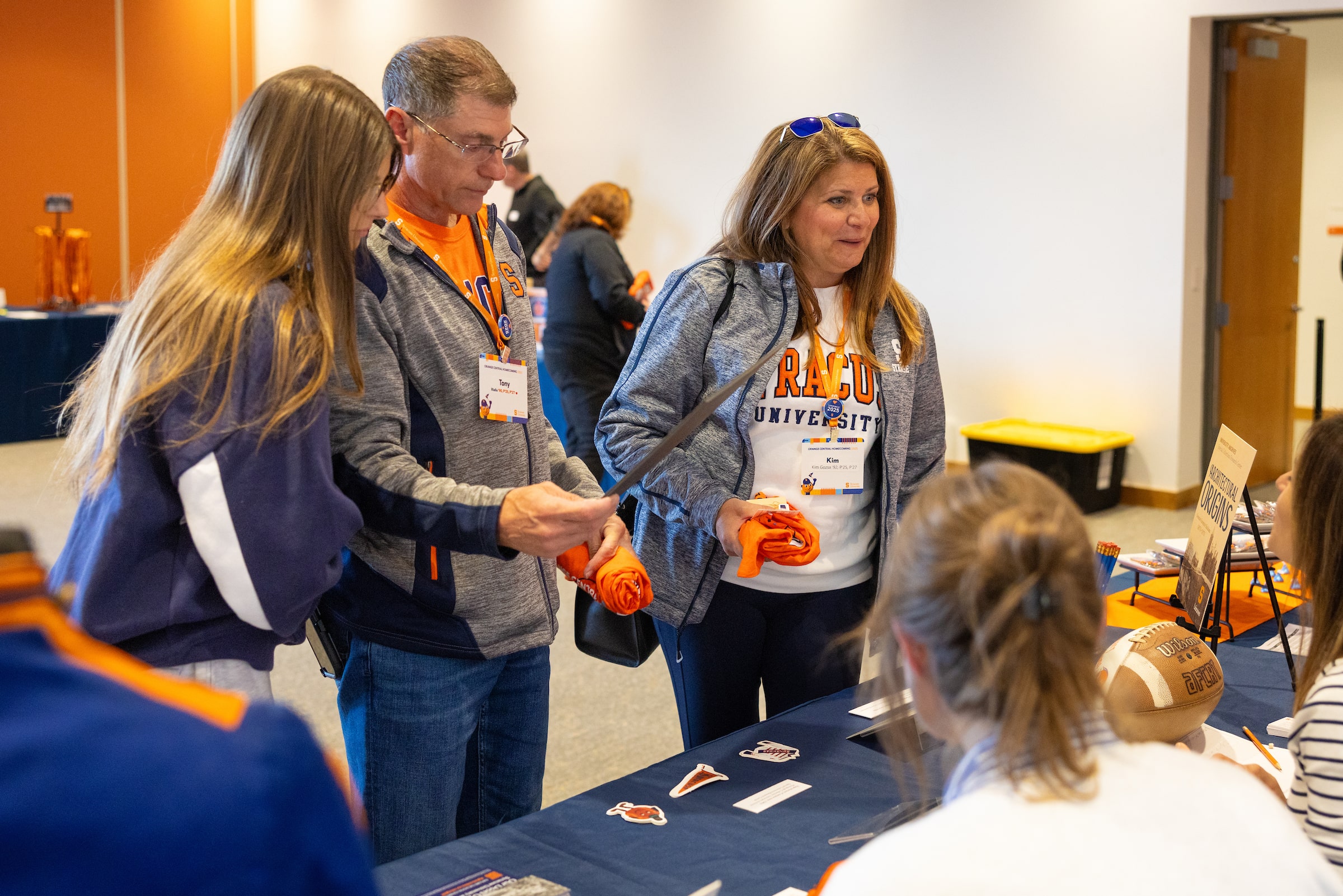 Three people gather at a registration table, holding orange rolled items while interacting with staff at a campus event.