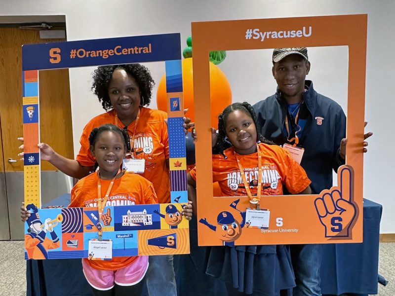 Family of Syracuse University fans posing with branded photo frames at a campus event, wearing orange "Orange Central Homecoming" shirts and displaying school spirit.
