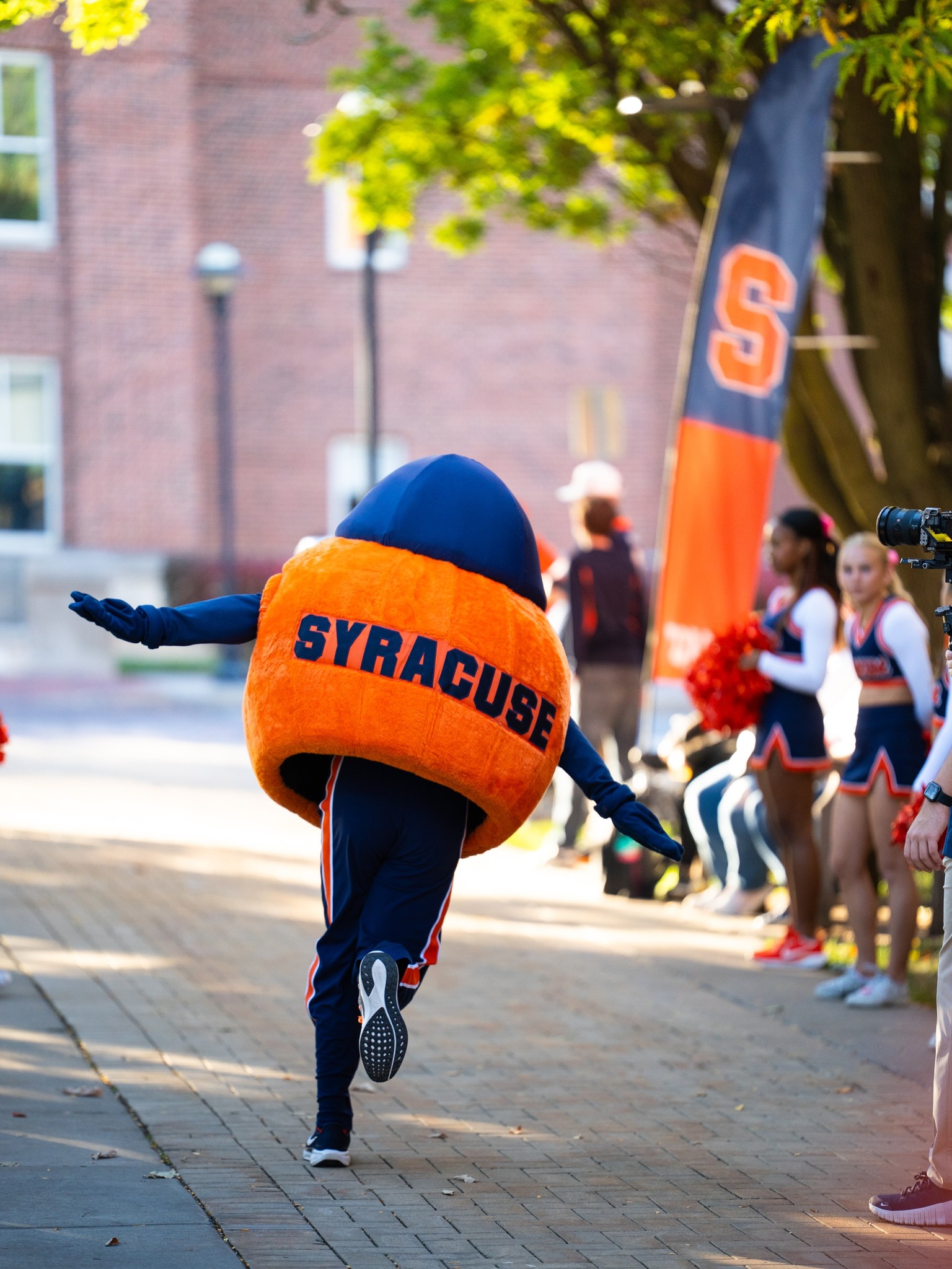 Otto the Orange mascot mid-stride on brick walkway with cheerleaders visible in background during outdoor event