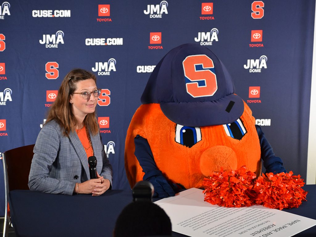 Otto the Orange with a woman holding a microphone at left. Otto is holding orange pom poms and a NIL deal document is on the table in front of him.