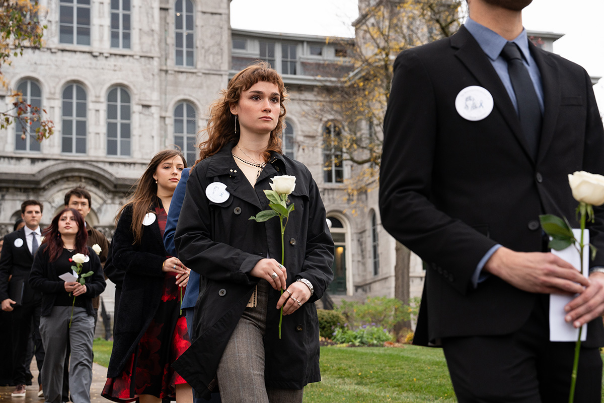 Group of formally dressed individuals walking in a line, each holding a white rose, during a commemorative event outside a historic stone building with arched windows