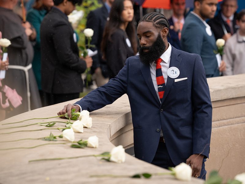 Person in navy suit placing a white rose on a curved stone memorial wall during a solemn commemorative event, with others holding roses in the background