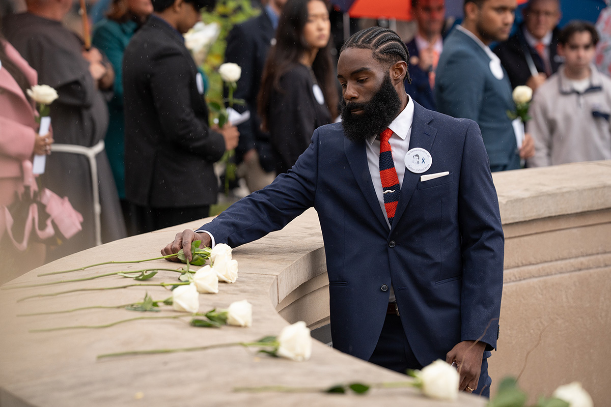 Person in navy suit placing a white rose on a curved stone memorial wall during a solemn commemorative event, with others holding roses in the background