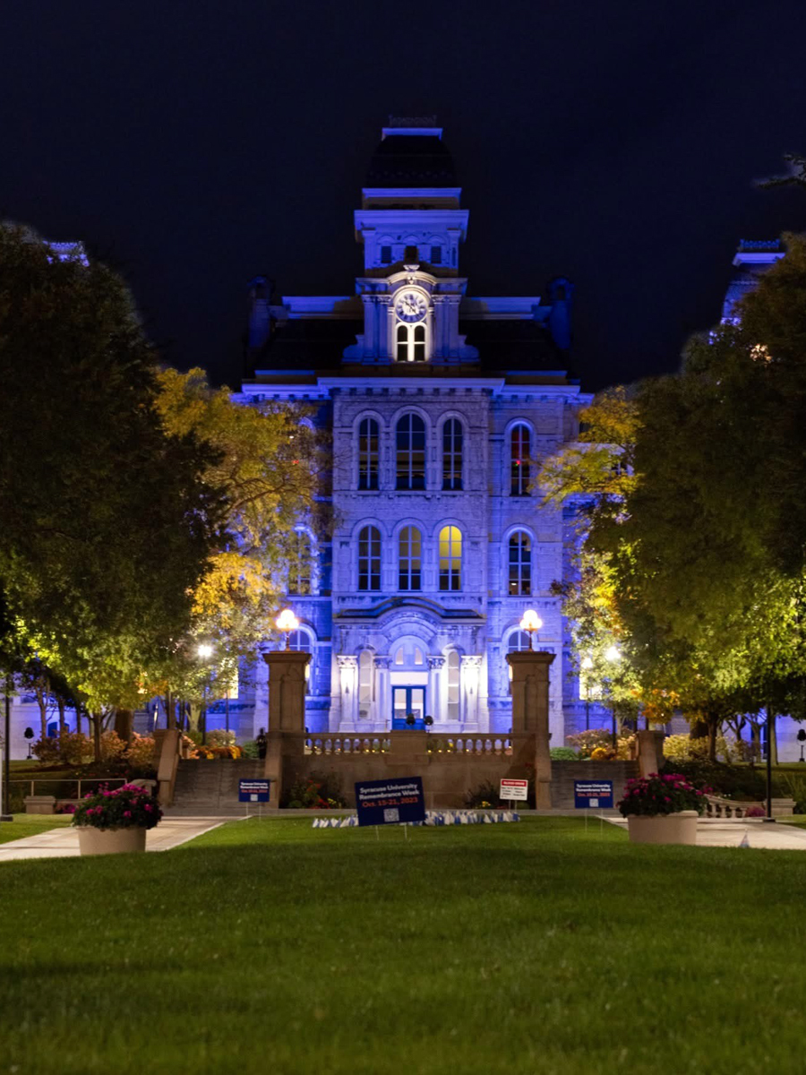 Historic university building illuminated in blue light at night, with a clock tower, ornate architecture, and tree-lined walkway leading to the entrance.