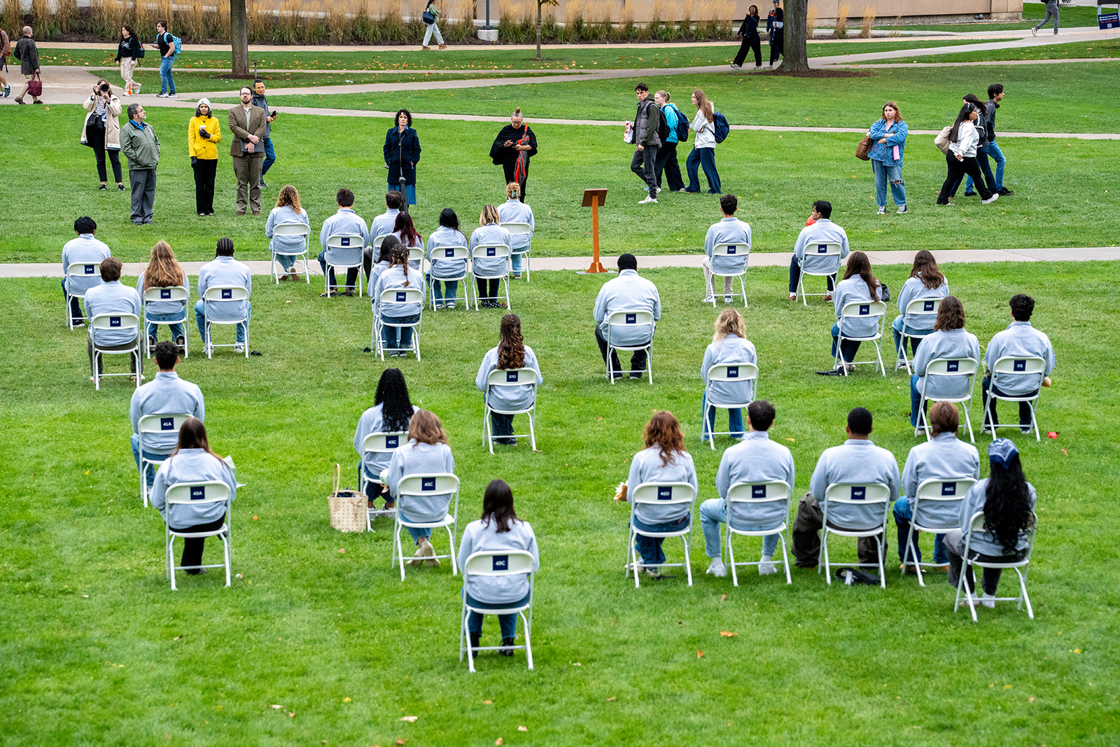 A large group of seated attendees in matching light jackets during an outdoor campus ceremony with white folding chairs and walking paths.
