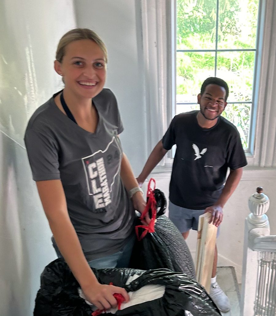 Two volunteers smile while standing on a stairway landing while holding large black garbage bags and pieces of wood.