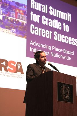A professor speaks at a podium with a presentation in the background displaying "Rural Summit for Cradle to Career Success: Advancing Place-Based Initiatives Nationwide"