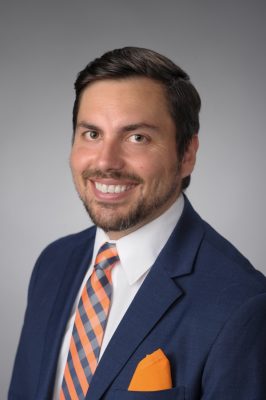 Person wearing a navy blue suit jacket with an orange pocket square, a white dress shirt, and an orange-and-navy striped tie