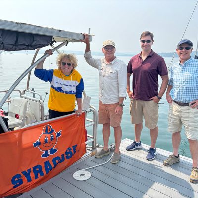 Group of people by a boat with a Syracuse University flag on a calm lake.