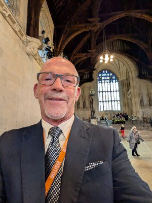 A man in a dark suit and patterned tie wearing an orange visitor lanyard takes a selfie inside Westminster Hall, with its iconic vaulted timber ceiling and Gothic architecture visible behind him. A large stained glass window and ornate stonework frame the historic interior space.