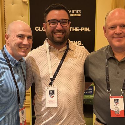 Three people standing indoors wearing name badges, with a “Syracuse Alumni” banner in the background.