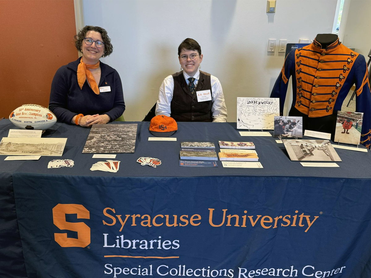 Syracuse University Libraries Special Collections staff at a display table featuring historic university memorabilia including a marching band uniform, vintage beanie, photographs, and archival documents.
