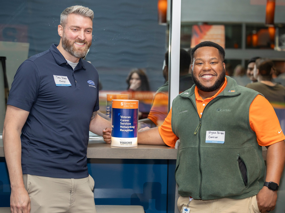 Two men standing at a reception table with a blue and orange banner for Syracuse University's Veteran Career Services Networking Reception, both smiling at the camera.