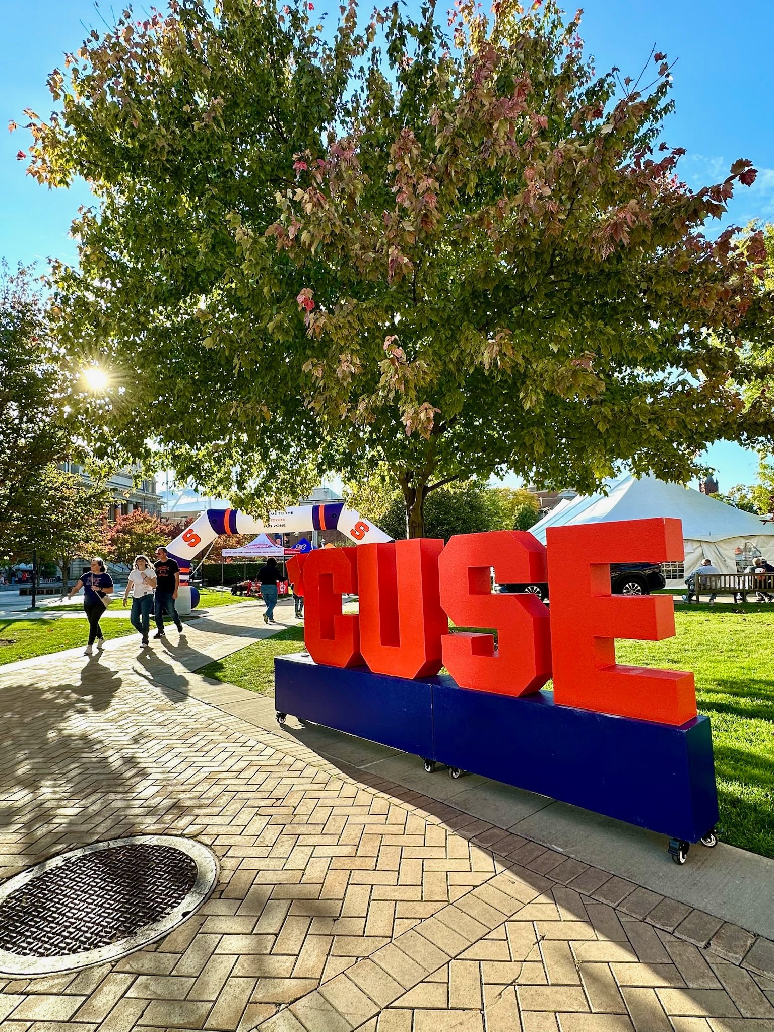Large three-dimensional "CUSE" letters in orange and blue displayed on campus quad under tree with event tents behind.