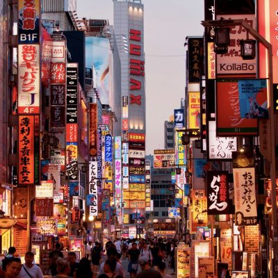 A bustling Tokyo street at dusk filled with colorful neon signs, crowded shops, and people walking among vibrant advertisements and restaurant lights.