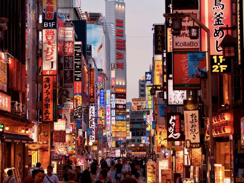 A bustling Tokyo street at dusk filled with colorful neon signs, crowded shops, and people walking among vibrant advertisements and restaurant lights.