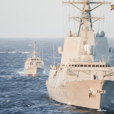 Two NATO naval ships sail in formation across open ocean waters during a joint maritime operation. The larger vessel in the foreground flies the flag of Spain, with another ship following behind under clear skies and moderate waves.