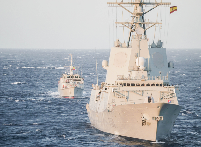 Two NATO naval ships sail in formation across open ocean waters during a joint maritime operation. The larger vessel in the foreground flies the flag of Spain, with another ship following behind under clear skies and moderate waves.