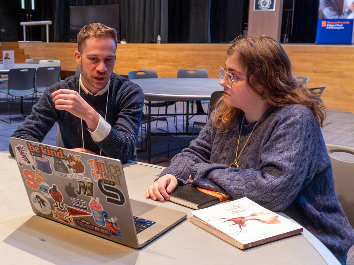 An alumnus chats with a current student at a table with laptop and textbooks in a large event space or auditorium.